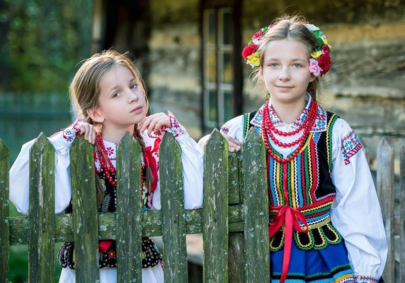 dwie dziewczynki w polskich strojach ludowych i koralach na szyi przy płocie